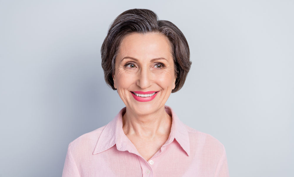 Charming elderly woman in pink smiling, representing seniors evaluating implants versus partial dentures.
